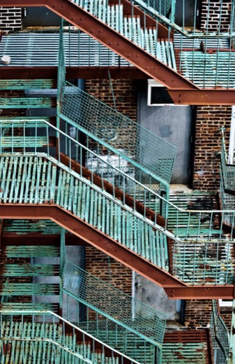 Architectural photo looking up at a weathered teal and rust colored metal fire escape staircase zigzagging down a brick building.