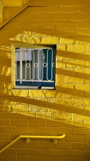 A bright yellow brick stairwell wall with a square cutout window showing a white railing and a teal interior beyond, lit by diagonal sunbeams.